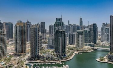 A high-angle aerial photograph of the Dubai Marina under a clear blue sky. The scene features a deep green canal filled with several white luxury yachts docked at piers.