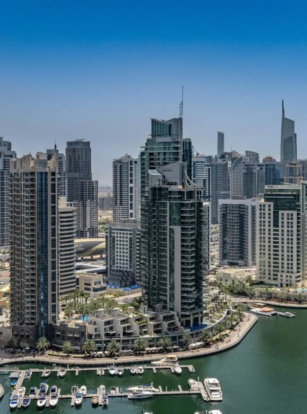A high-angle aerial photograph of the Dubai Marina under a clear blue sky. The scene features a deep green canal filled with several white luxury yachts docked at piers.