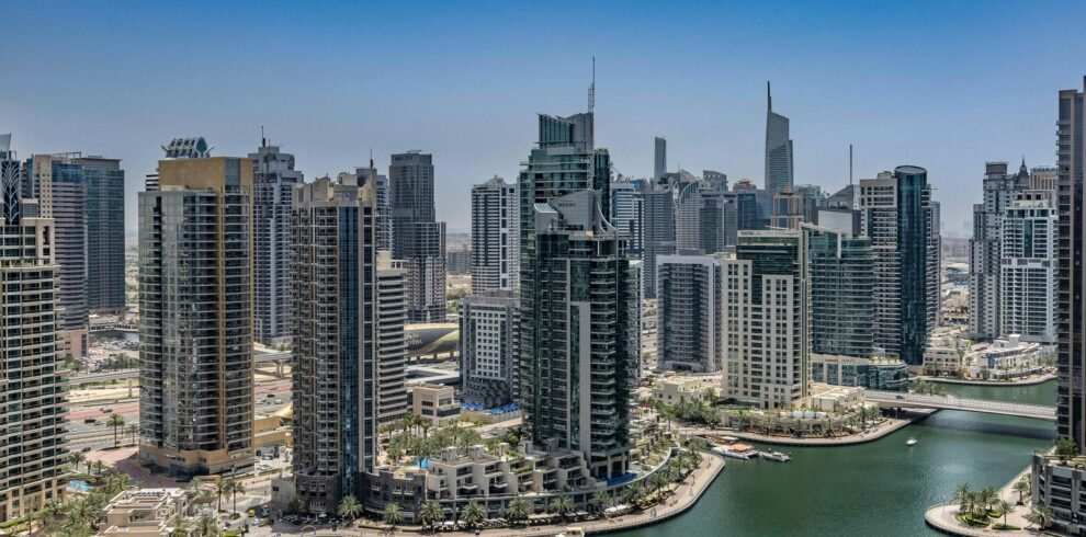 A high-angle aerial photograph of the Dubai Marina under a clear blue sky. The scene features a deep green canal filled with several white luxury yachts docked at piers.