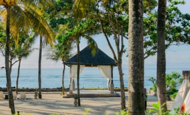 A serene view of a traditional wooden gazebo with a dark thatched roof and white flowing curtains on a white sand beach.