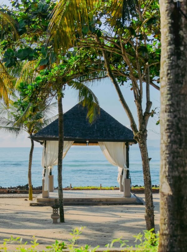 A serene view of a traditional wooden gazebo with a dark thatched roof and white flowing curtains on a white sand beach.