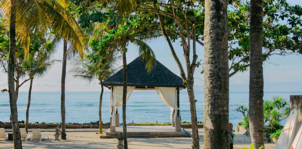 A serene view of a traditional wooden gazebo with a dark thatched roof and white flowing curtains on a white sand beach.