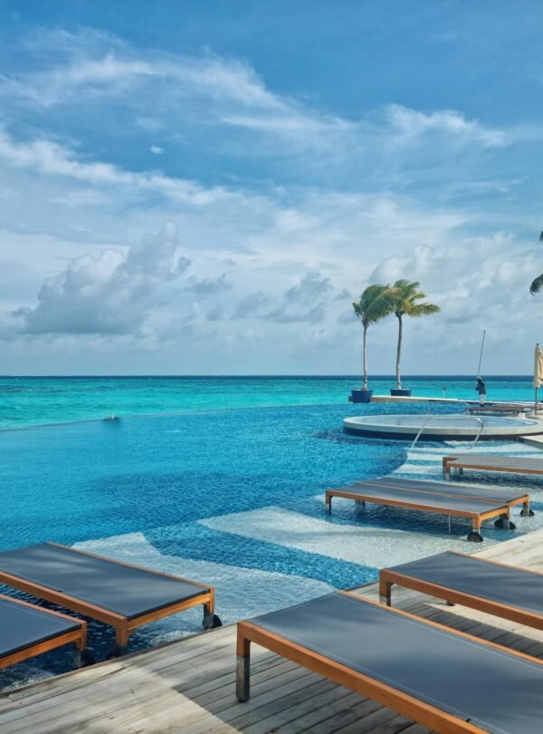 A wide-angle view of a curved infinity pool overlooking a turquoise ocean under a bright, cloudy sky.