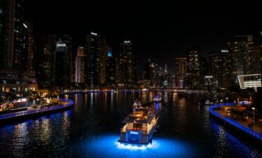 A high-angle night view of a luxury yacht with bright blue underwater lights cruising through the Dubai Marina canal.