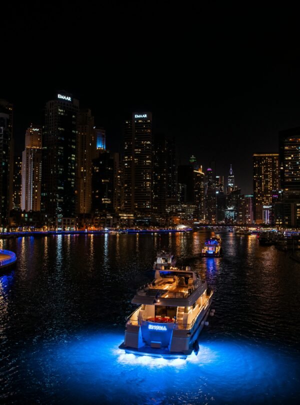 A high-angle night view of a luxury yacht with bright blue underwater lights cruising through the Dubai Marina canal.
