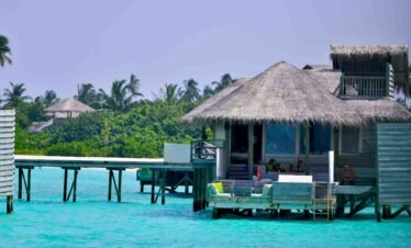 A close-up view of a luxury overwater villa with a thatched roof and light-colored wooden walls built on stilts over a vibrant turquoise lagoon.