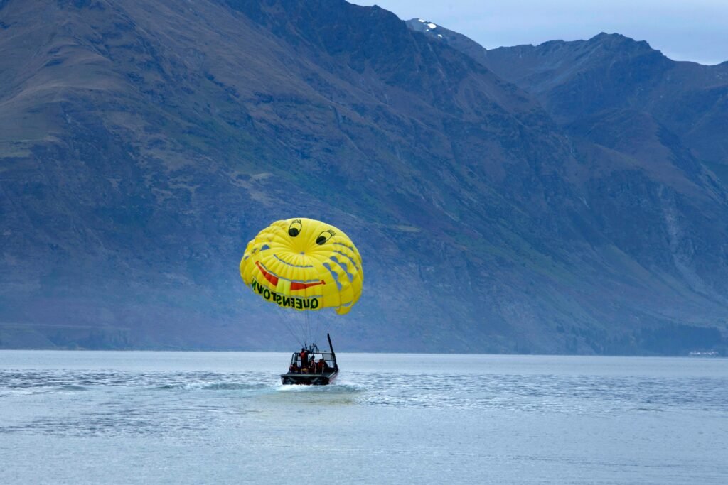 A yellow smiley-face parachute is towed by a black boat across a calm lake, with the steep, rugged, and dark blue mountains of Queenstown, New Zealand, rising in the background under an overcast sky.