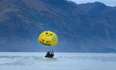 A yellow smiley-face parachute is towed by a black boat across a calm lake, with the steep, rugged, and dark blue mountains of Queenstown, New Zealand, rising in the background under an overcast sky.