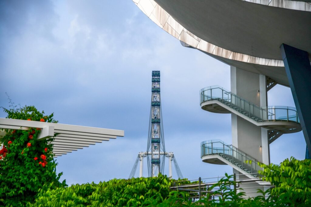 A vertical side-on view of the Singapore Flyer observation wheel framed by the modern white architecture and curved glass-railed balconies of Gardens by the Bay. In the foreground, lush green tropical plants and vibrant red flowers climb a white trellis under a cloudy blue sky.